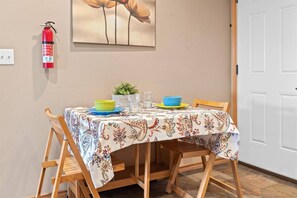 Une table à manger recouverte d’une nappe à motifs floraux, avec des chaises en bois et des bols colorés. Un extincteur est fixé au mur, au-dessus de la table.