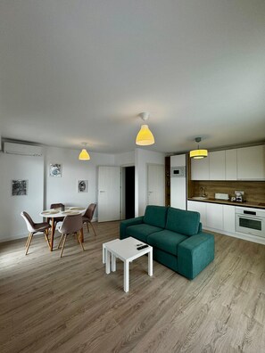 Una sala de estar moderna con un sofá verde, una mesa de comedor con sillas, y un área de cocina con armarios blancos y una encimera de madera.
