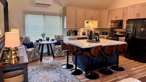 Una cocina moderna con armarios de madera, una isla central con taburetes de bar, un área de comedor con sofás estampados y un ventanal con persianas.