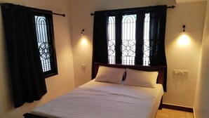 A neatly made bed with a wooden headboard, two windows with decorative iron grilles, and wall-mounted lights. - The Chill House (Kochi)