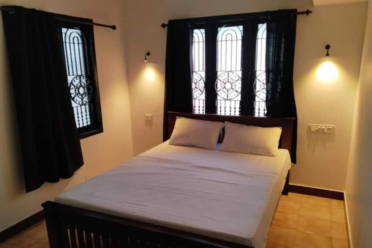 A neatly made bed with a wooden headboard, two windows with decorative iron grilles, and wall-mounted lights.