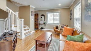 A spacious living room with hardwood floors, a wooden coffee table, and a brown leather sofa with orange cushions. A staircase with white railings leads to another room.
