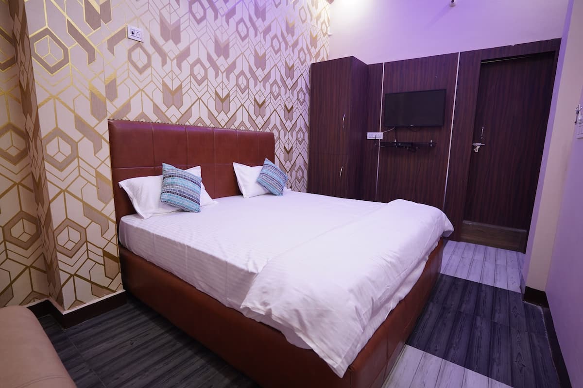 A neatly made bed with white bedding and decorative pillows, a wooden wardrobe with a television, and patterned wallpaper.