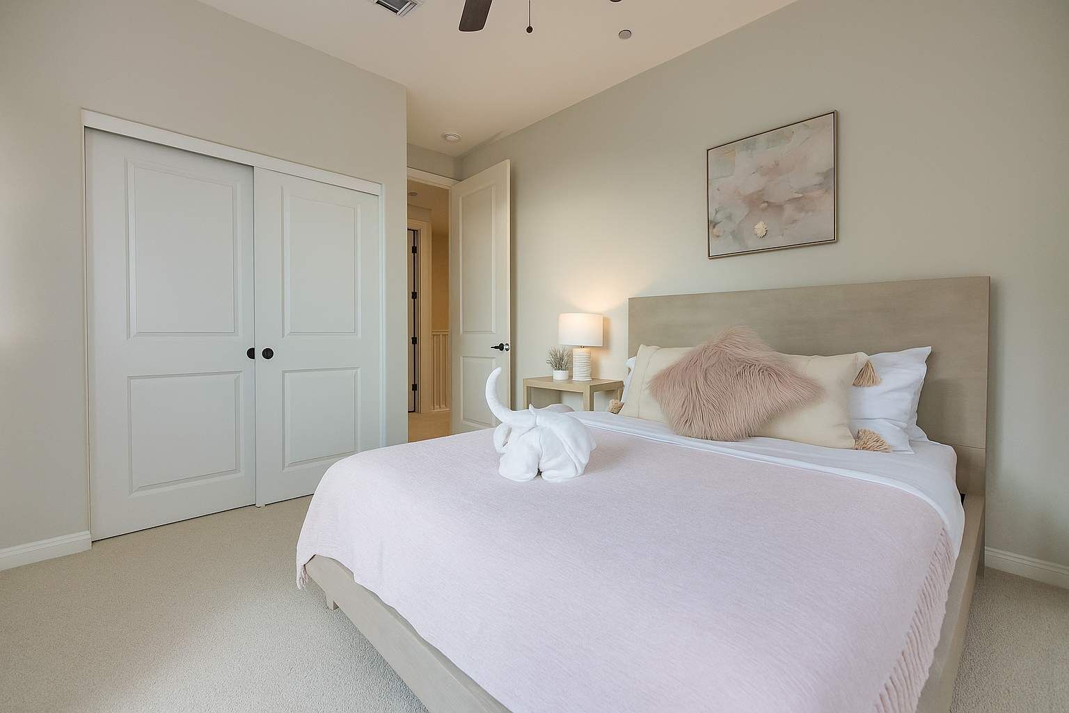 A bedroom with a large bed, a nightstand with a lamp, a painting on the wall, and a door leading to another room.