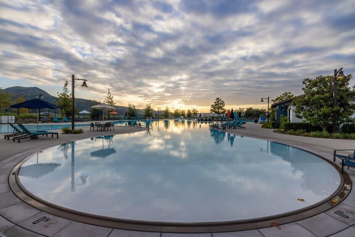 Una gran piscina circular con sillas de descanso y sombrillas, rodeada de árboles y edificios, bajo un cielo nublado.