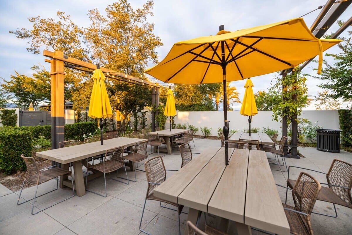 An outdoor patio area with wooden tables and chairs, yellow umbrellas, and a pergola structure.