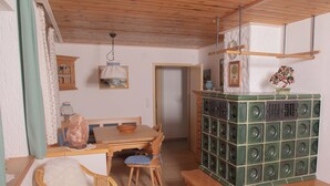 Een gezellige kamer met een houten plafond, een eettafel met stoelen, een groene kast met speakers, en een rieten fauteuil met een zacht kussen.