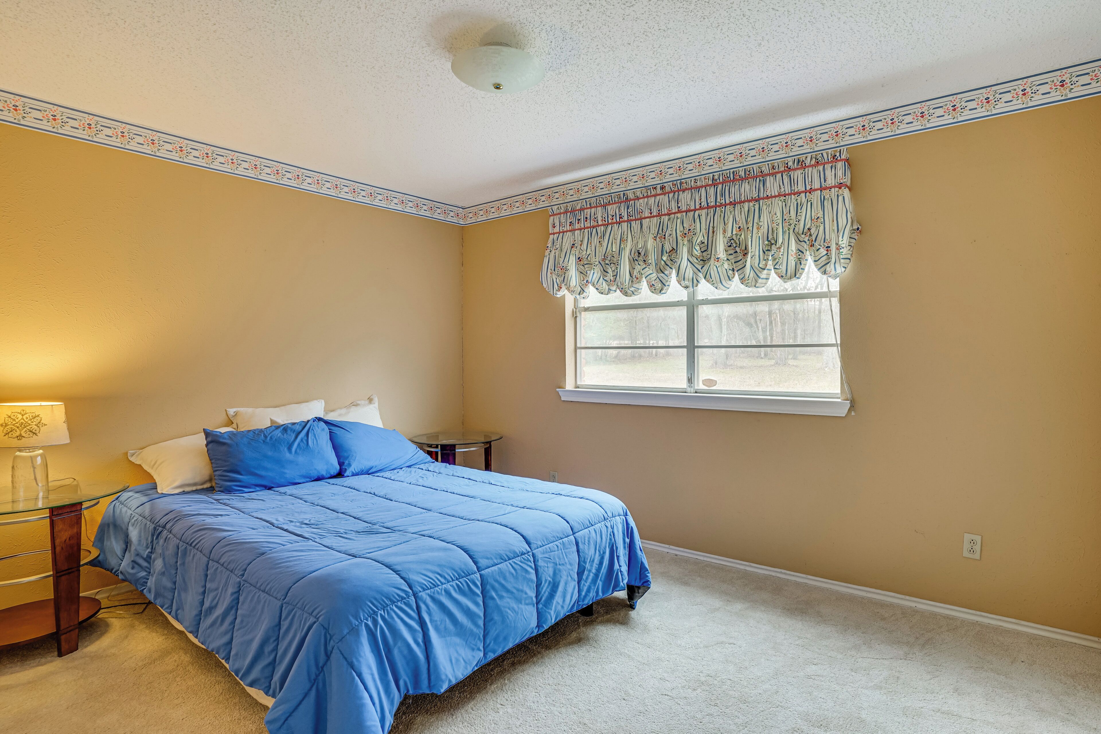 Une chambre à coucher comprenant un lit, une table de chevet, une lampe, une fenêtre avec un lambrequin et un sol recouvert d’une moquette.