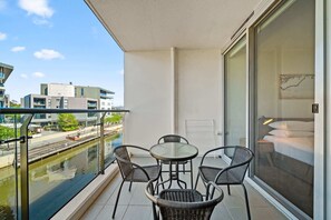 A balcony with a table and chairs, a view of a canal, and modern buildings in the background.
