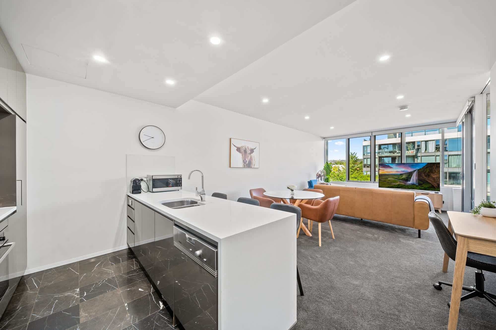 A modern kitchen with a sink, microwave, and clock, a dining area with chairs and a table, and a large window with a view of buildings.