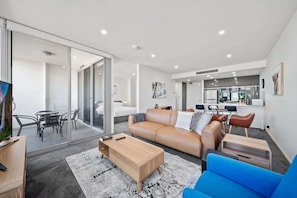 A modern living room with a sofa, coffee table, and blue armchair. There is a dining area with a table and chairs, and a kitchen in the background.