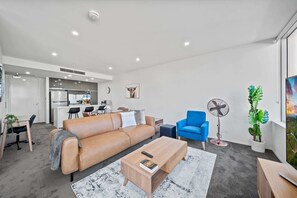 A modern living room with a sofa, coffee table, and blue armchair. There is a kitchen in the background and a large window with a plant.