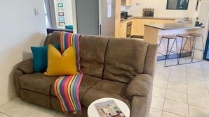 Een woonkamer met een bruine bank, een kleurrijke plaid en een kleine ronde tafel met een tijdschrift. In de achtergrond is een keuken met een koelkast en een bar te zien.