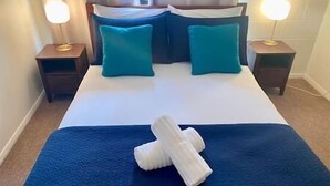 Een net opgemaakt bed met een blauwe spreideken en twee teal kussens, aan weerszijden geflankeerd door twee nachtkastjes met lampen.
