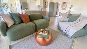 Een moderne woonkamer met een groene bank, een ronde salontafel en een eethoek op de achtergrond.