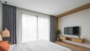 A modern hotel room with a large bed, a flat-screen TV mounted on the wall, and a large window with sheer curtains.