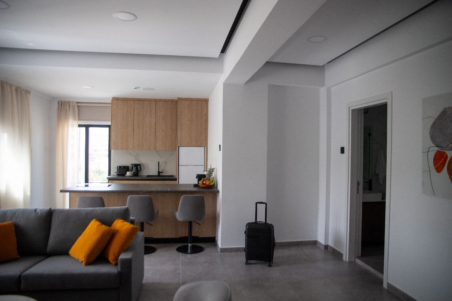 A modern hotel room with a grey sofa, a small kitchen area, and a suitcase on the floor.