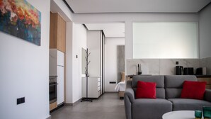 A modern living room with a grey sofa, red cushions, a small round table, and a kitchen area in the background. - LE BOUQUET LUXURY SUITES  (Nicosia)