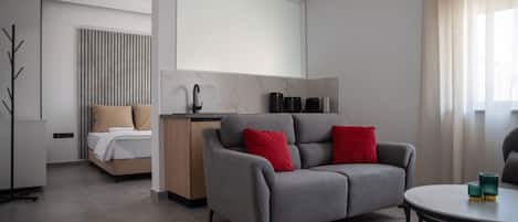 Ein modernes Hotelzimmer mit einer grauen Couch, einem Bett mit beige- und roten Kissen, einem kleinen Tisch mit Gläsern und einem großen Fenster mit durchsichtigen Vorhängen.