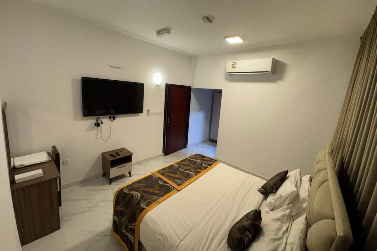 Quarto de hotel com cama, televisão na parede, ar condicionado, escrivaninha com abajur e guarda-roupa.
