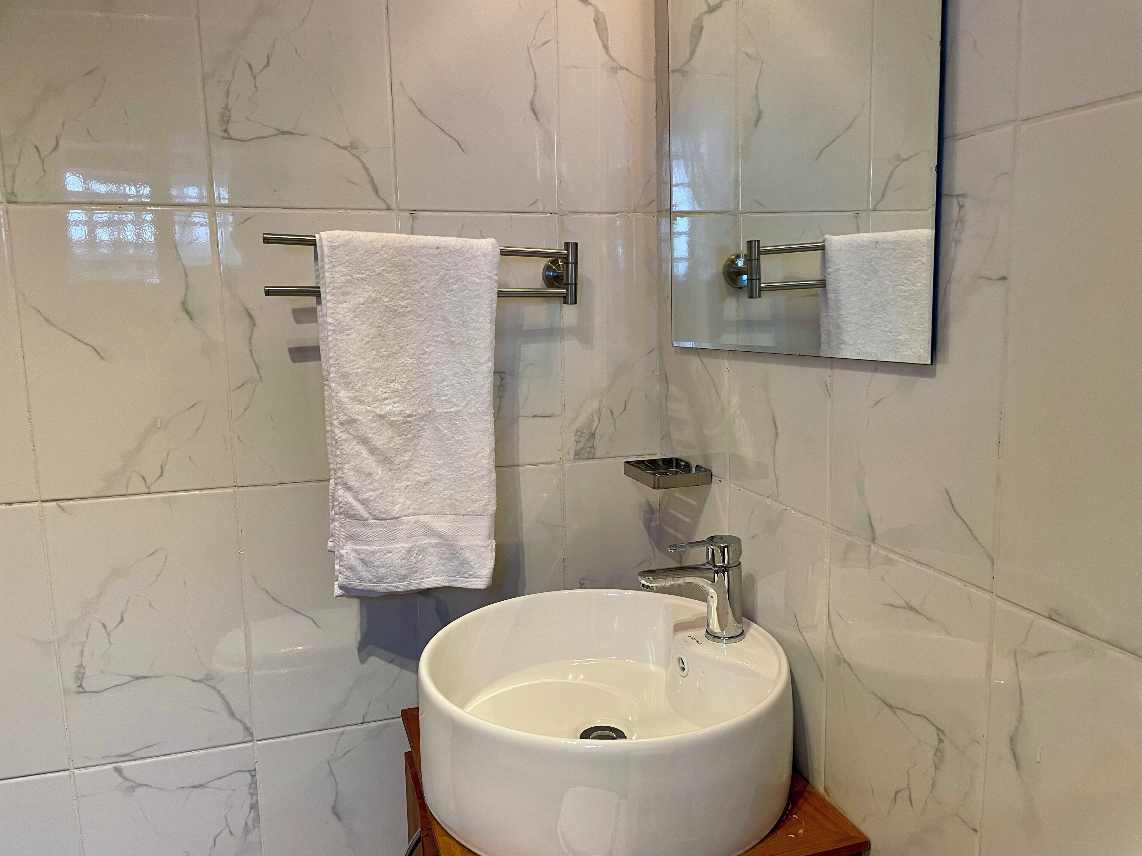 Un baño con pared de blanco mármol, un lavamanos blanco con grifo cromado y un espejo sobre el lavamanos.