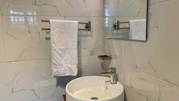 Un baño con pared de blanco mármol, un lavamanos blanco con grifo cromado y un espejo sobre el lavamanos.