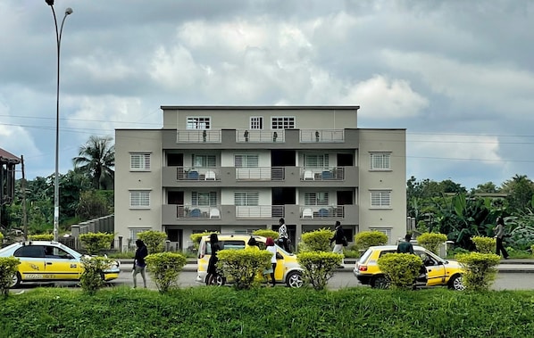 A multi-story residential building with balconies, a parking area with yellow cars, and greenery in the foreground.