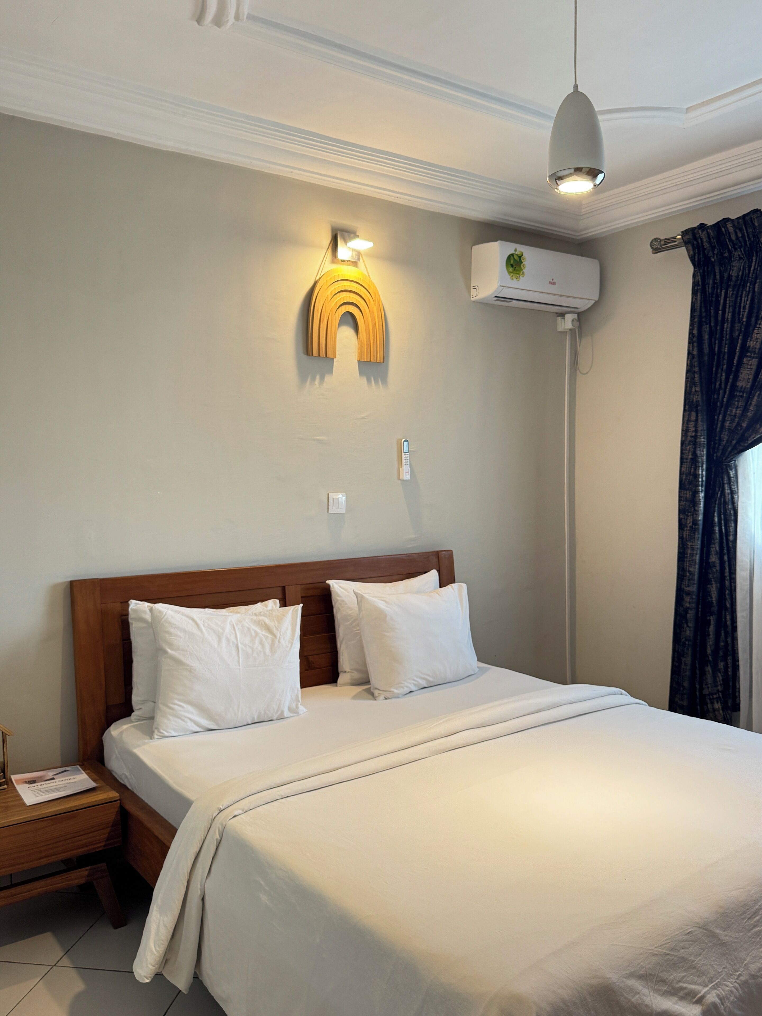 A hotel room with a wooden headboard, a bed with white linens, a wall-mounted air conditioning unit, and a pendant light.