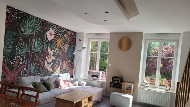A modern living room with a large tropical-themed wall tapestry, a wooden coffee table, a grey sofa, and a white ottoman. - Spacious duplex at Matthieu's. Sleeps 1 to 6. (Remiremont)