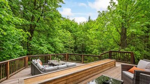 Une terrasse en bois dotée d'un canapé intégré et de meubles en osier, entourée d'arbres verdoyants.