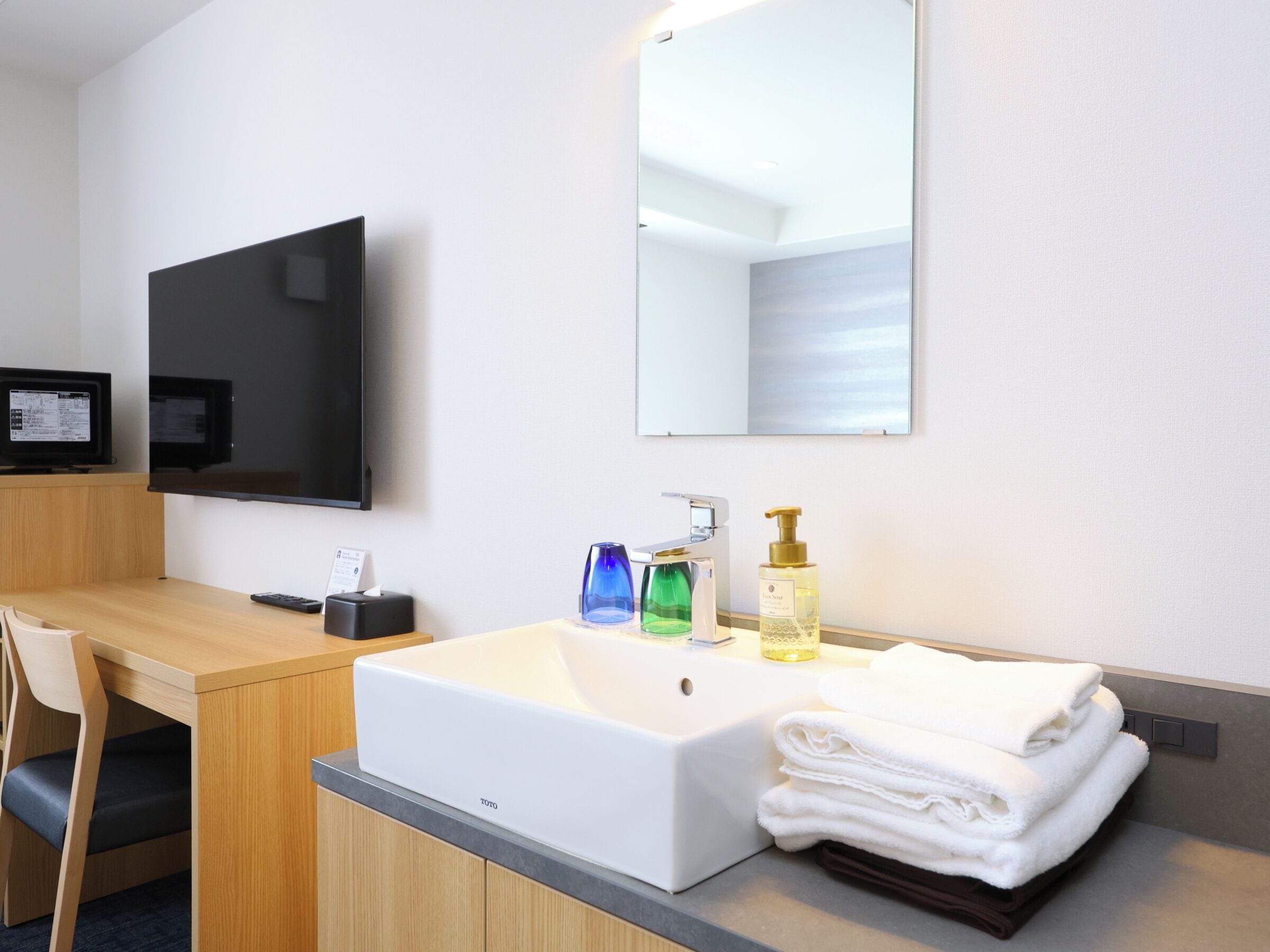 A hotel bathroom with a white sink, a mirror, a television, and a towel stack.