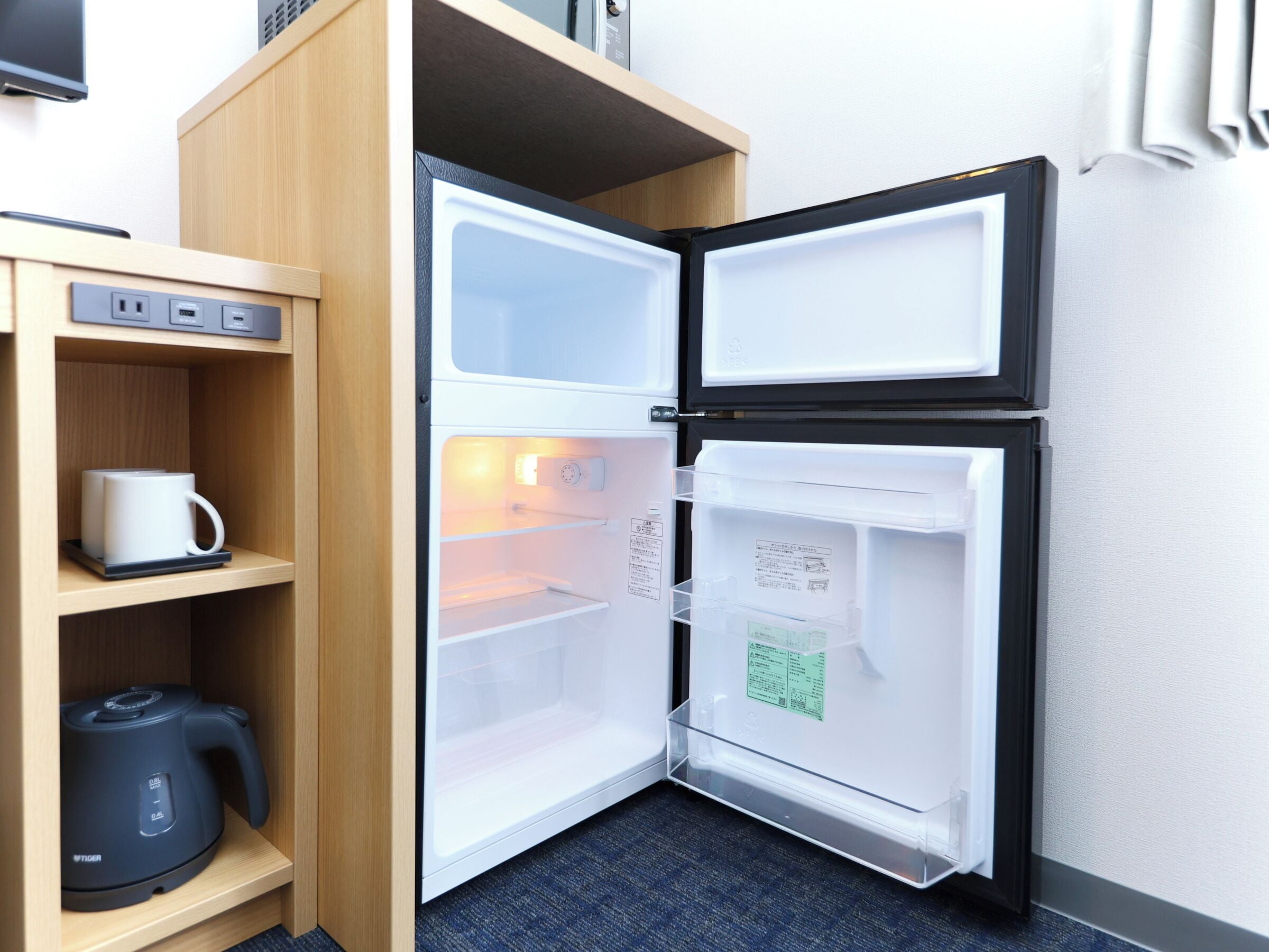 A compact refrigerator with a built-in coffee maker and a kettle, placed in a corner with a wooden shelf and a power outlet.