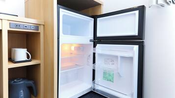 A compact refrigerator with a built-in coffee maker and a kettle, placed in a corner with a wooden shelf and a power outlet.