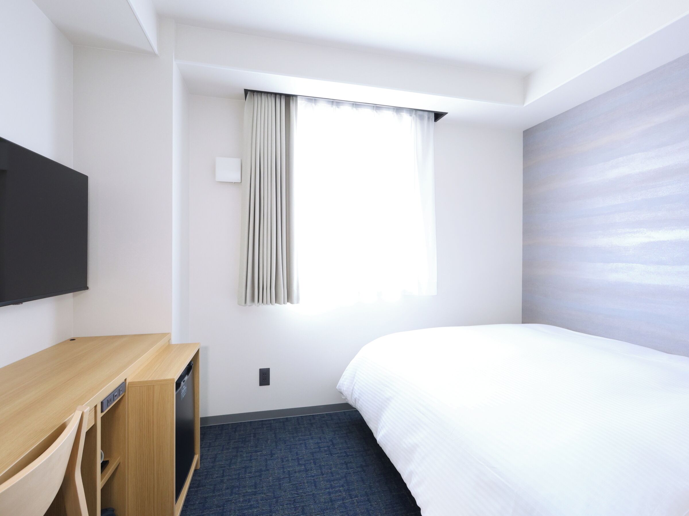 A hotel room with a bed, a desk with a television, a window with curtains, and a wall with a textured design.