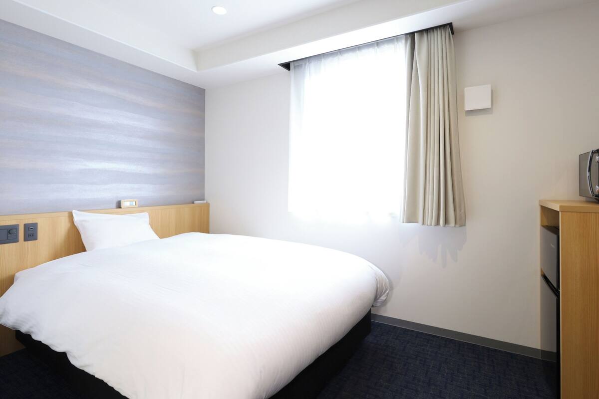 A hotel room with a bed, a window with curtains, a wall-mounted TV, and a small wooden cabinet.