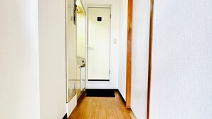 A narrow hallway with wooden flooring, a white door at the end, and a shoe rack with slippers at the bottom. - [ 303 ] Twin Japanese-style room with kitchen, bathroom and washing machine, 2 min. from the nearest station, along the airport line. (大阪市)