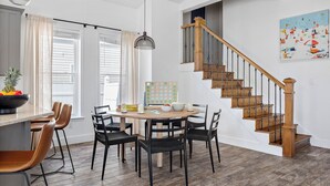 A dining area with a wooden table, black chairs, and a fruit bowl. A staircase with wooden steps and metal railings is visible.