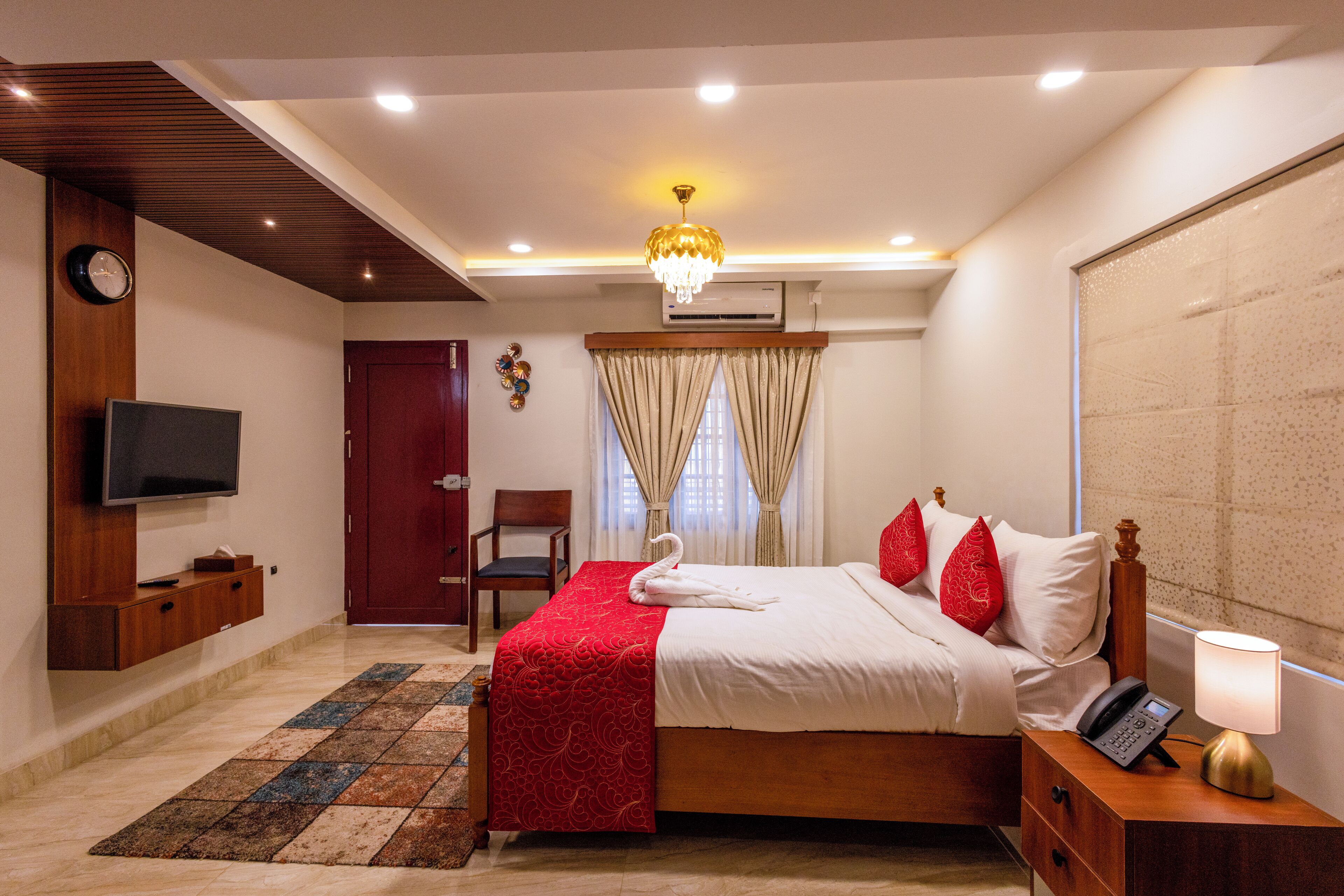 A hotel room with a bed, a television, a desk with a lamp, a chair, a clock, and a window with curtains.