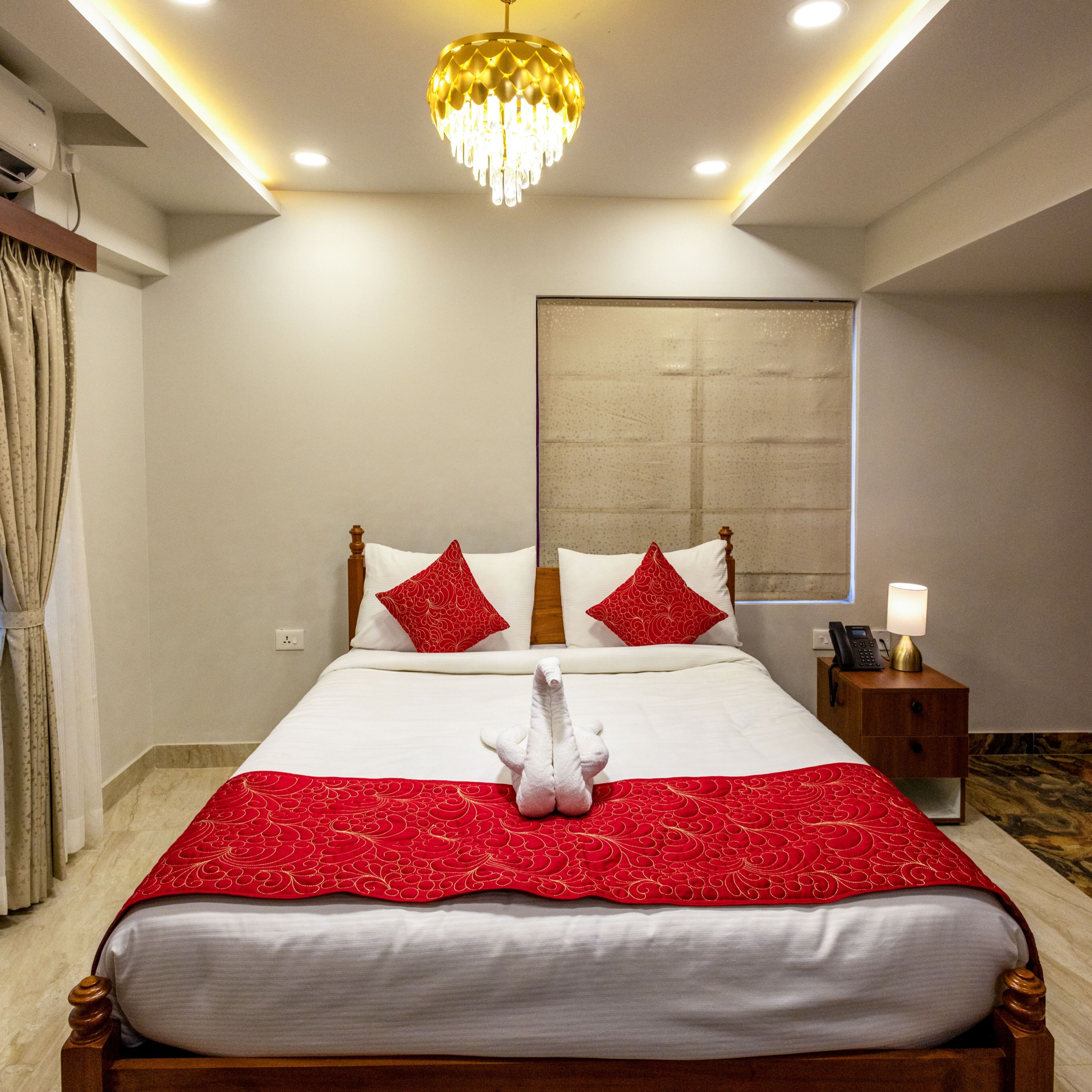 Una cama bien hecha con una colcha roja y blanca, dos almohadas rojas y un almohadón blanco y mullido. Se aprecia una mesita de noche de madera con una lámpara y un teléfono, una ventana grande con vistas, y un candelabro sobre la cama.