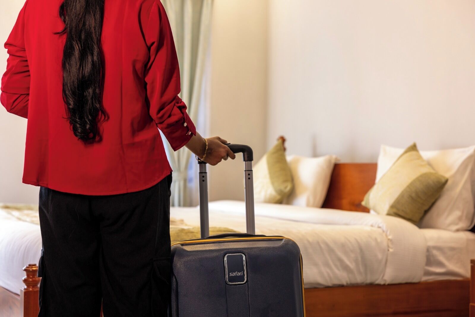 A person in a red shirt pulling a suitcase with a "safari" tag, standing in a hotel room with a bed and pillows.