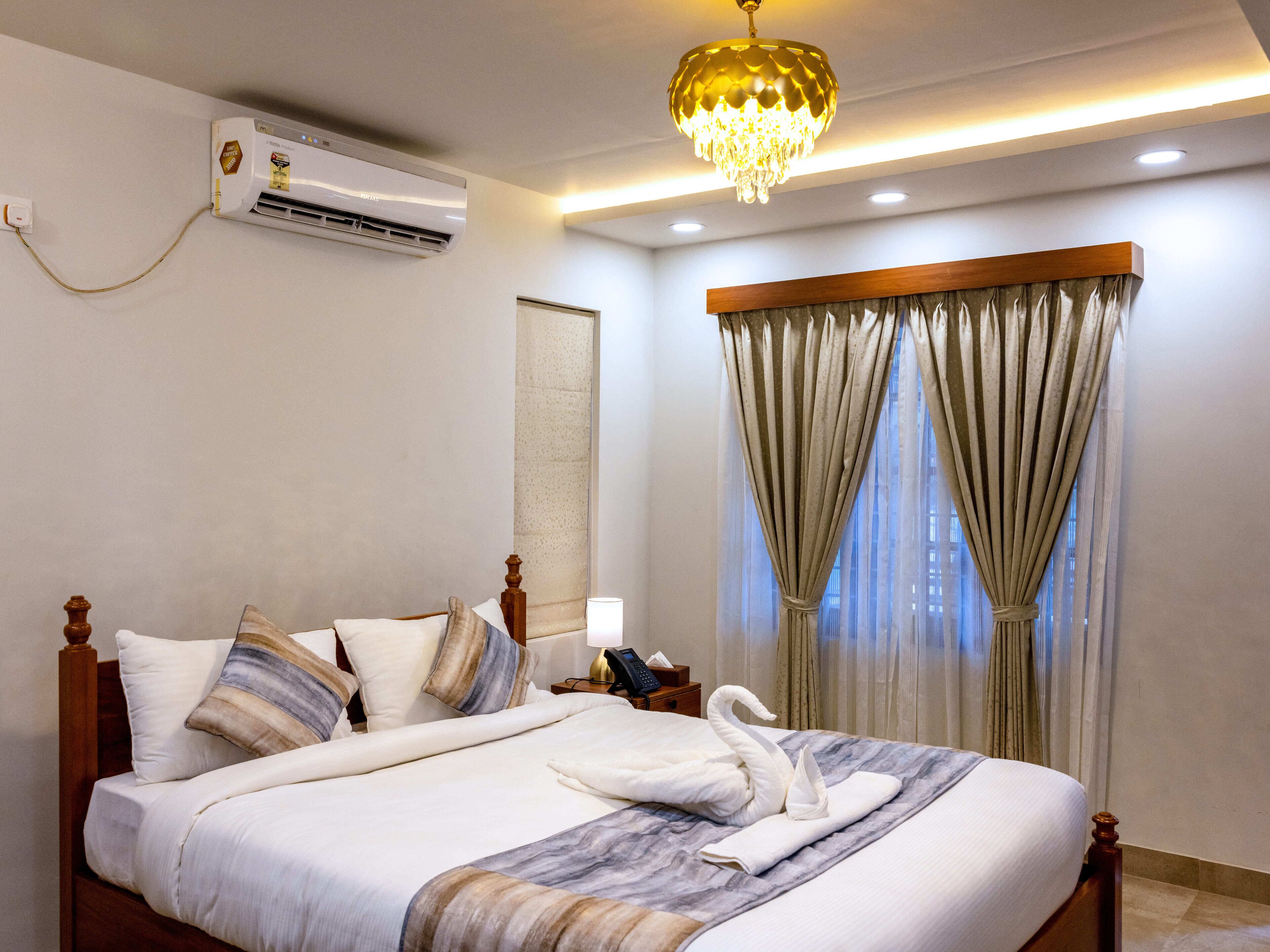 Une chambre d’hôtel avec un lit, une table de chevet, un appareil de climatisation, un lustre et une fenêtre avec des rideaux.