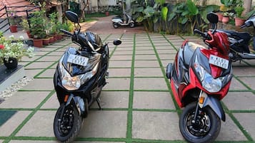 Two motorcycles parked on a paved area with a grid pattern, surrounded by potted plants and a small garden.