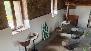 Un intérieur rustique avec des murs en pierre, un plafond en bois et un mélange de meubles modernes et traditionnels. On y trouve deux fenêtres, un banc en pierre et une chaise en osier noir.