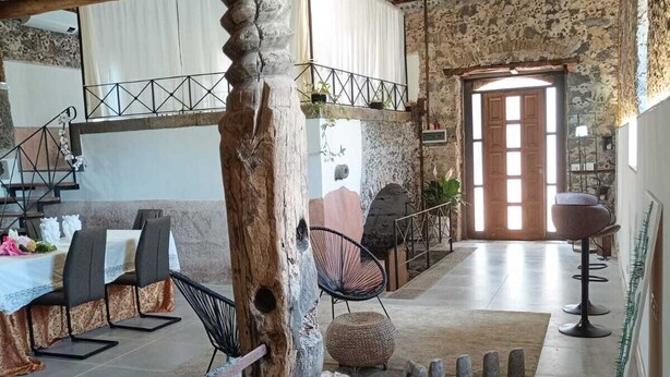 Un intérieur rustique avec un pilier en pierre, des poutres en bois, un coin repas avec des chaises et une table, et une vue sur l’extérieur par une porte.