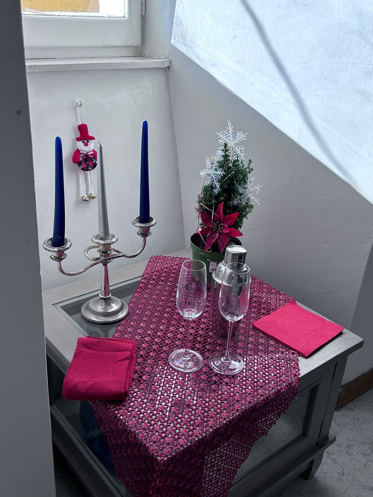 A small table with a pink tablecloth, two wine glasses, a candle holder with candles, a small Christmas tree, and a decorative snowflake.