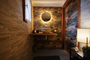 A bathroom with a wooden wall, a round mirror, a large bowl sink, a small trash can, and a nightstand with a lamp. - Relais Marmore with a Jacuzzi for two, a wood-burning stove in the room, and pets allowed (Ferentillo)
