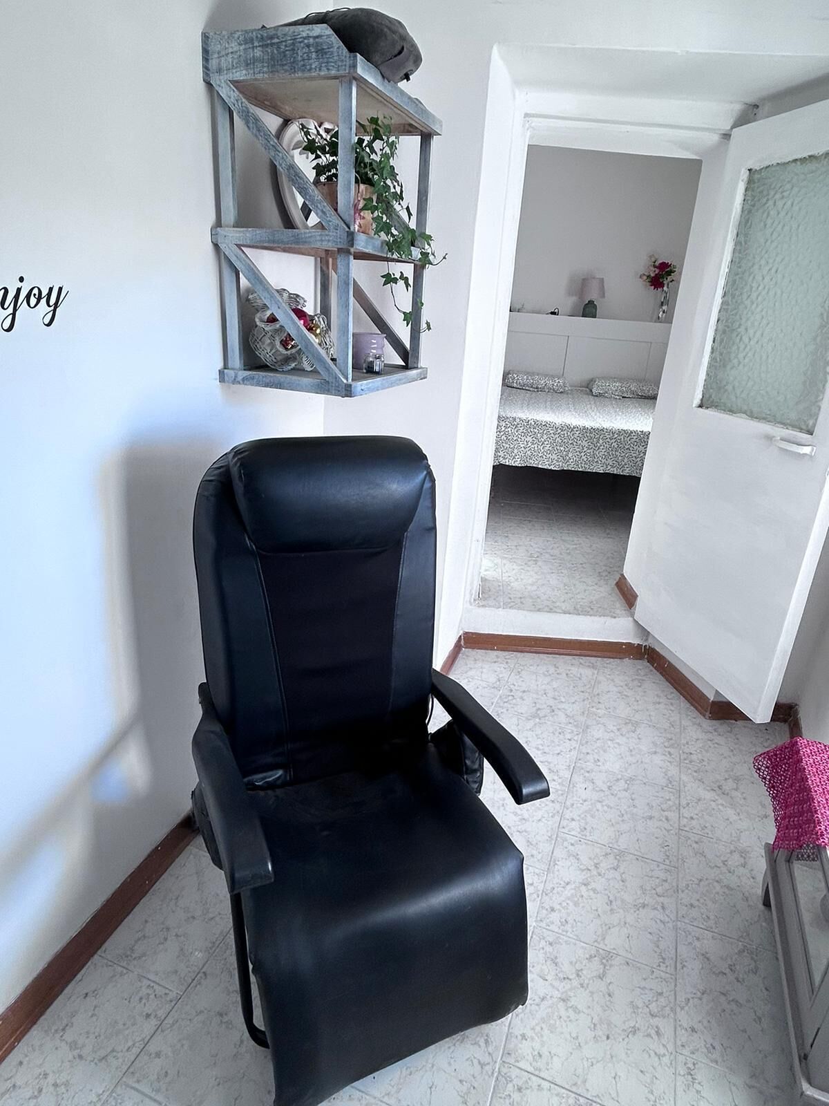 Una sedia da ufficio nera in una stanza con una porta bianca, una piccola mensola con oggetti decorativi e un letto con copripiumino fantasia.