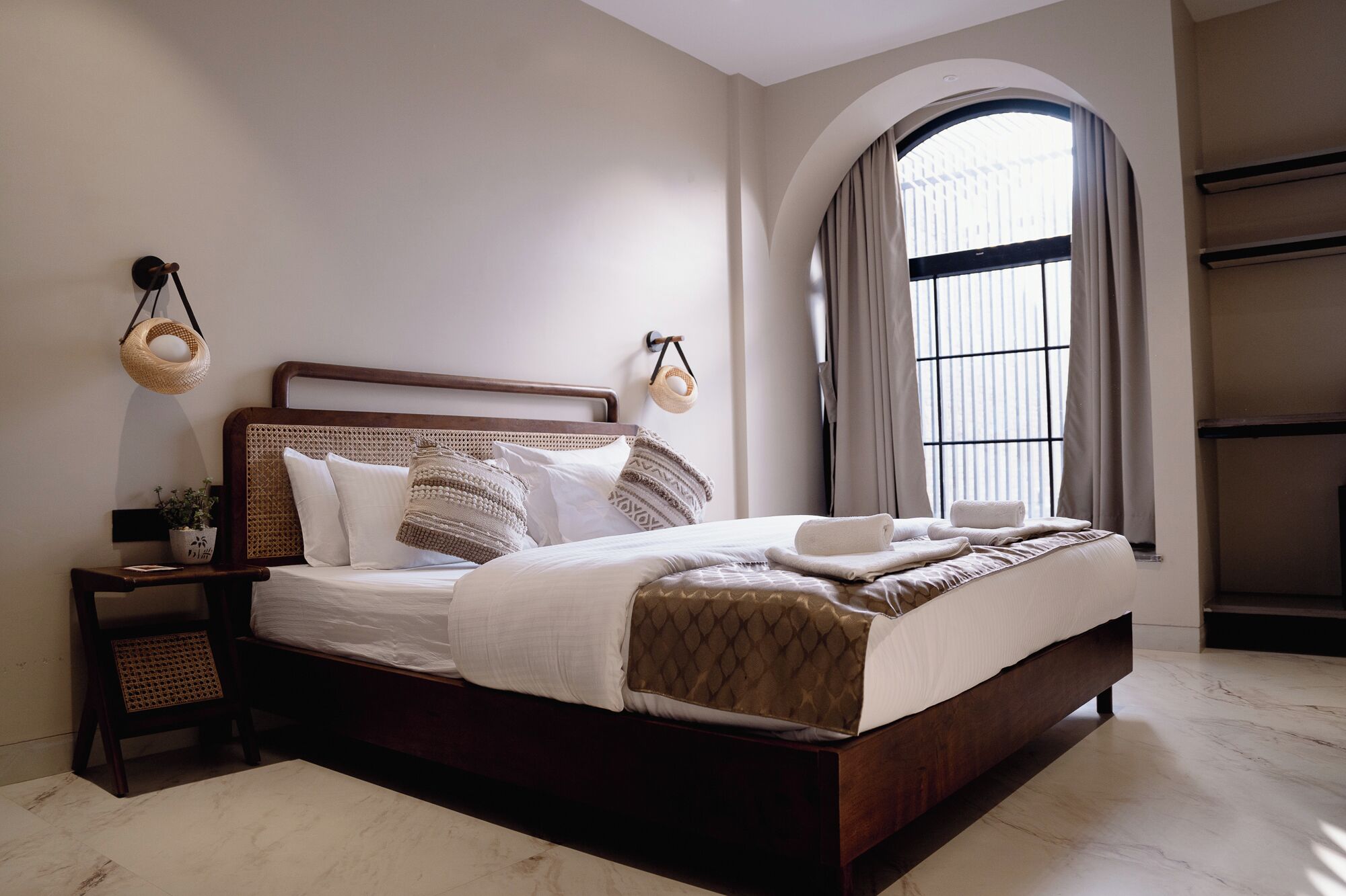 A neatly made bed with a wooden headboard, a bedside table with a lamp, and a large arched window with curtains.