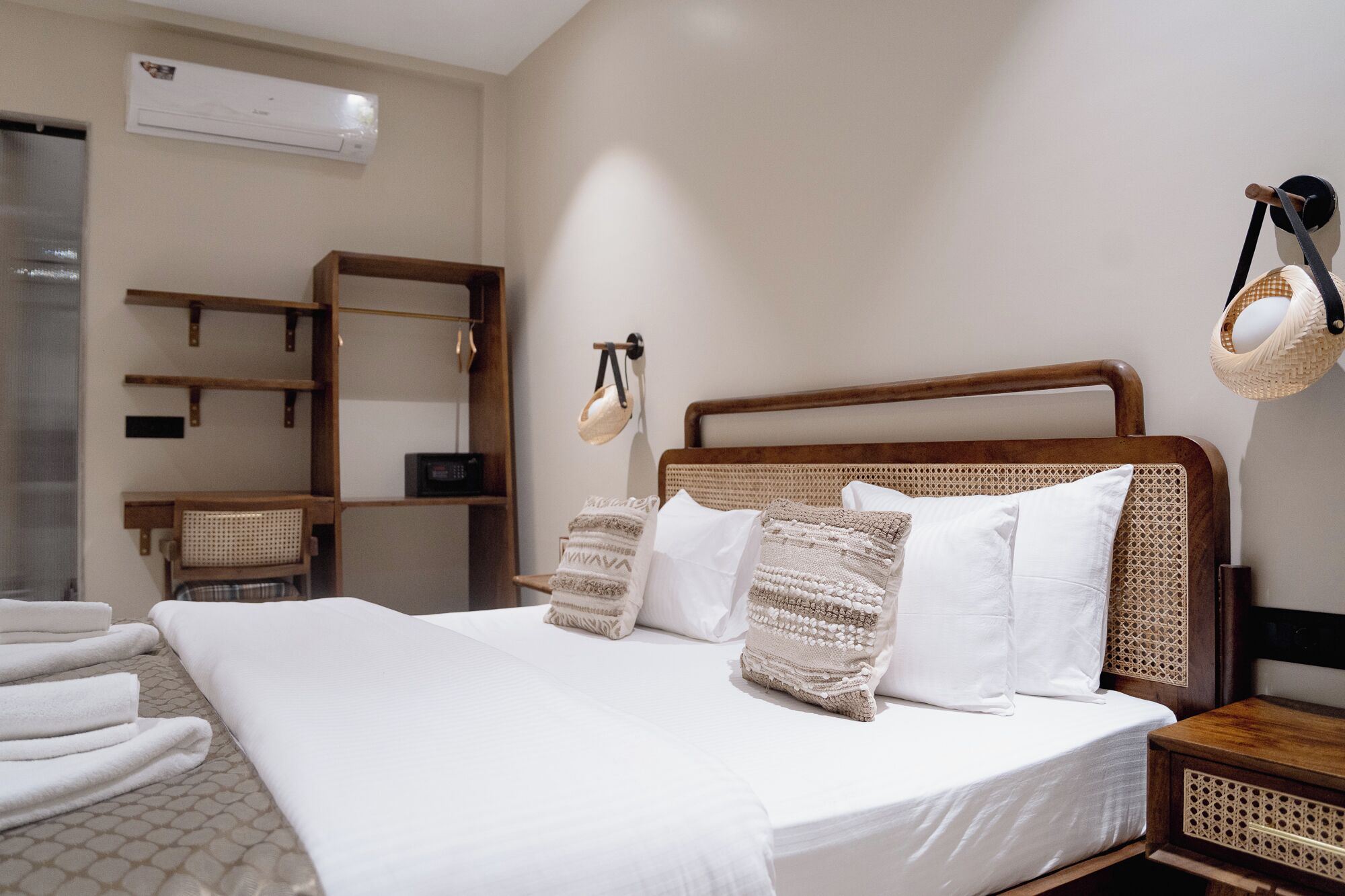 A neatly made bed with white linens and a woven headboard, a wooden nightstand with a woven basket, and a wall-mounted shelf with a woven basket and a small radio.
