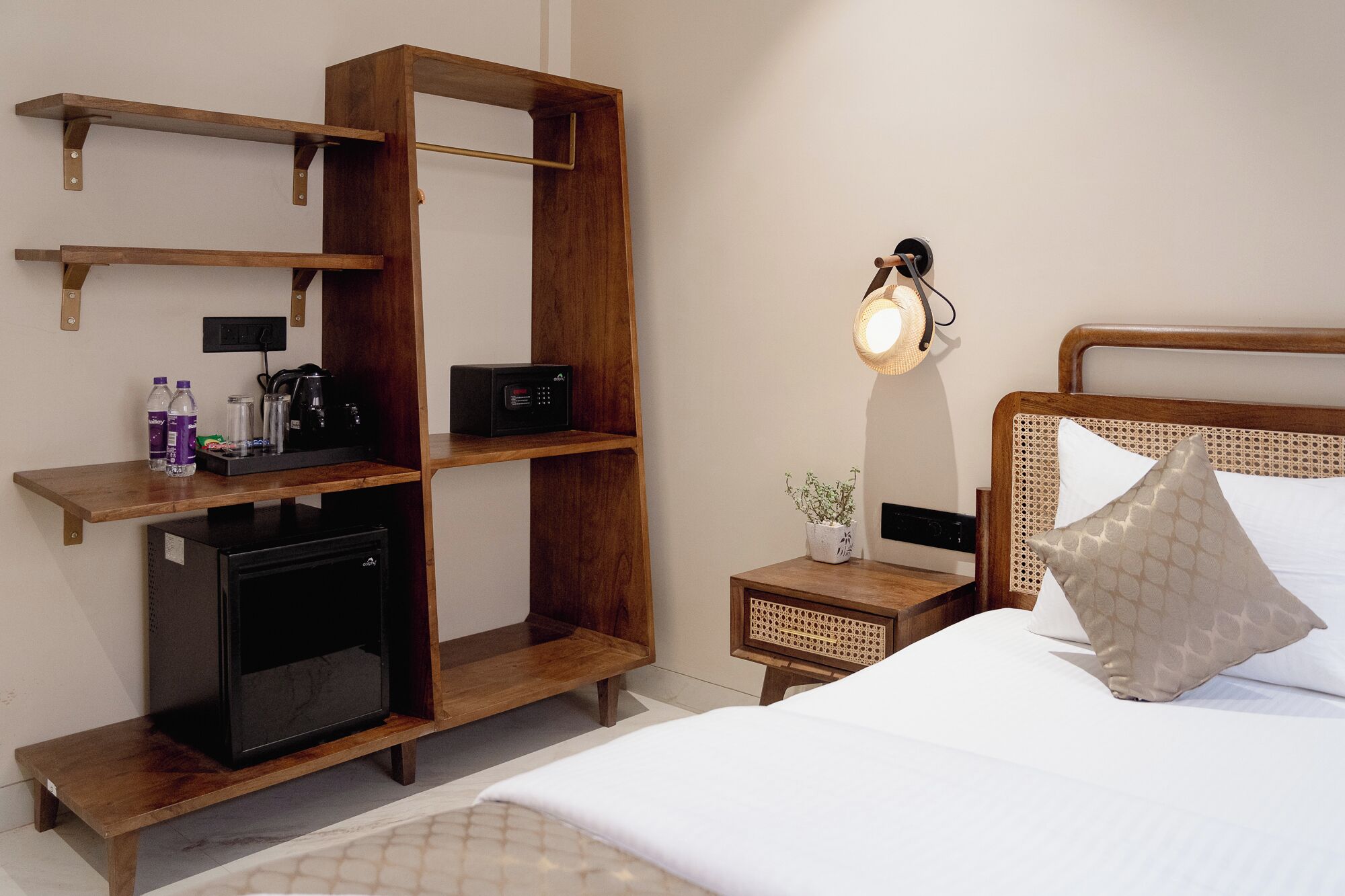 Quarto de hotel com cama, mesa de cabeceira, estante, micro-ondas, mini-geladeira, luminária e planta em vaso.
